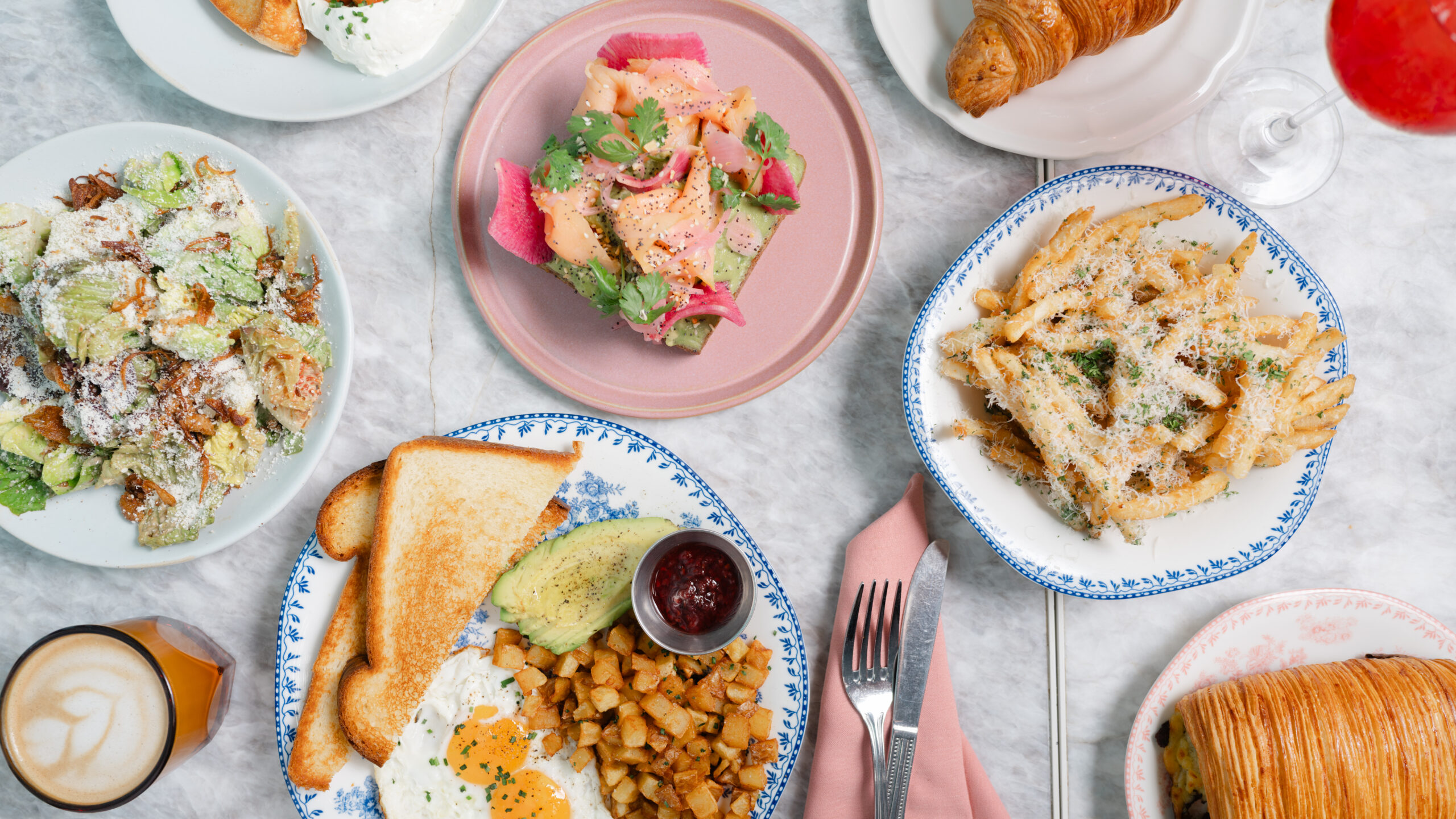 Oh La La – brunch spread of salad, pasta, eggs with toast, croissants and a latte on a marble table.