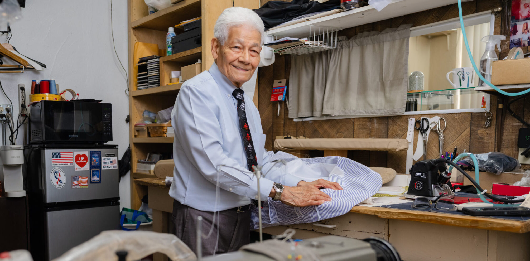 Noe Montoya Academy Tailor Shop – tailor working at his sewing bench surrounded by tools and garments.