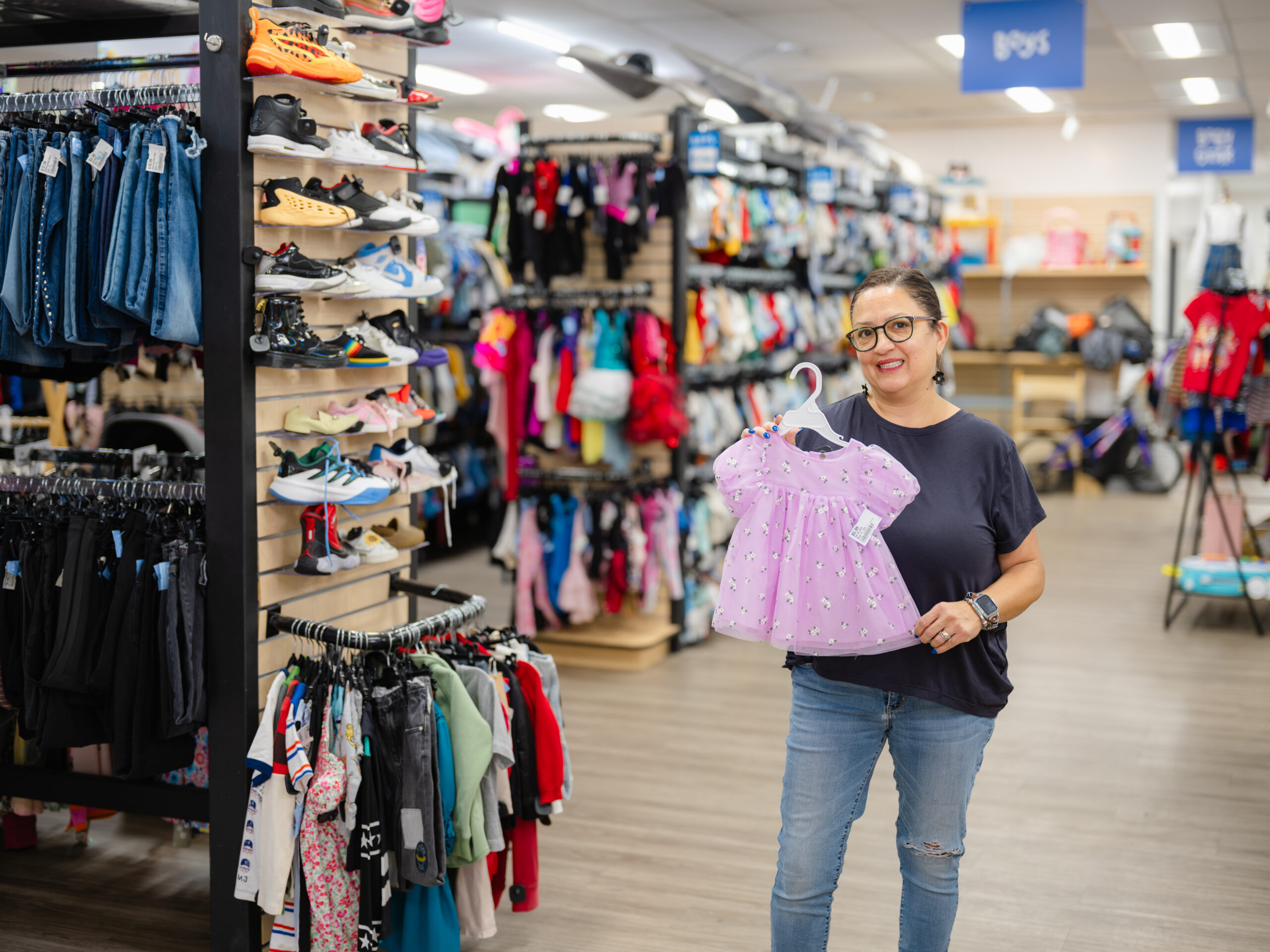 Kid to Kid – store associate holding a pink toddler dress among racks of children's clothing and shoes.